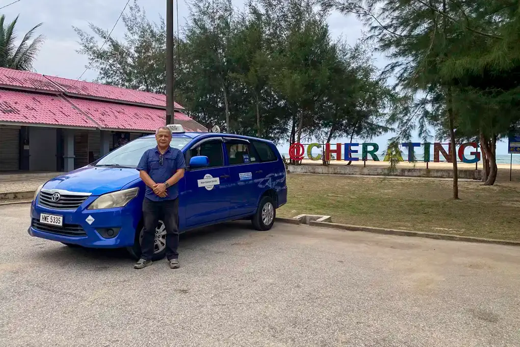 Anuar standing beside a blue Toyota Innova taxi near the Cherating beach sign