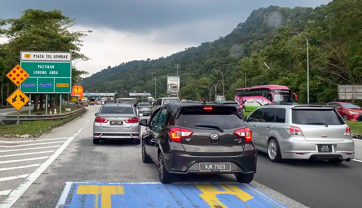 Heavy weekend traffic approaching Plaza Tol Gombak on the route out of Kuala Lumpur, with vehicles queueing below forested hills.